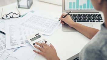 Persona calculando datos financieros y de nómina con documentos y gráficos, simbolizando la revisión contable y fiscal en una oficina.