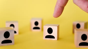 Imagen de un dedo de primer plano señalando un cubo de madera con una figura humana, simbolizando la selección y auditoría de proveedores de RH y operativos de servicios especializados en México bajo el cumplimiento normativo REPSE y mitigación de responsabilidad solidaria, sobre un fondo amarillo.